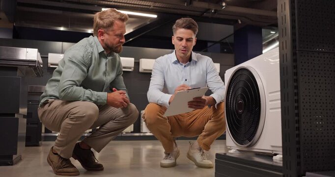 Salesman and customer consultation in hvac store. They assess an airconditioner as advisor notes specs on a clipboard to guide appliance choosing and budget. Clear retail guidance and decision.