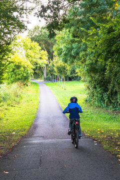 Young boy riding a bike on a park path lined with green trees and a sunlit lawn in summer, vertical rear view of a child cyclist in a blue jacket and helmet on a paved trail, concept of childhood, cy