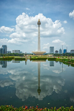 Indonesia, Jakarta &ndash; December 12, 2025: Monas tower reflected in a wide pool with green lawns and modern buildings, seen from Merdeka Square under a cloudy bright