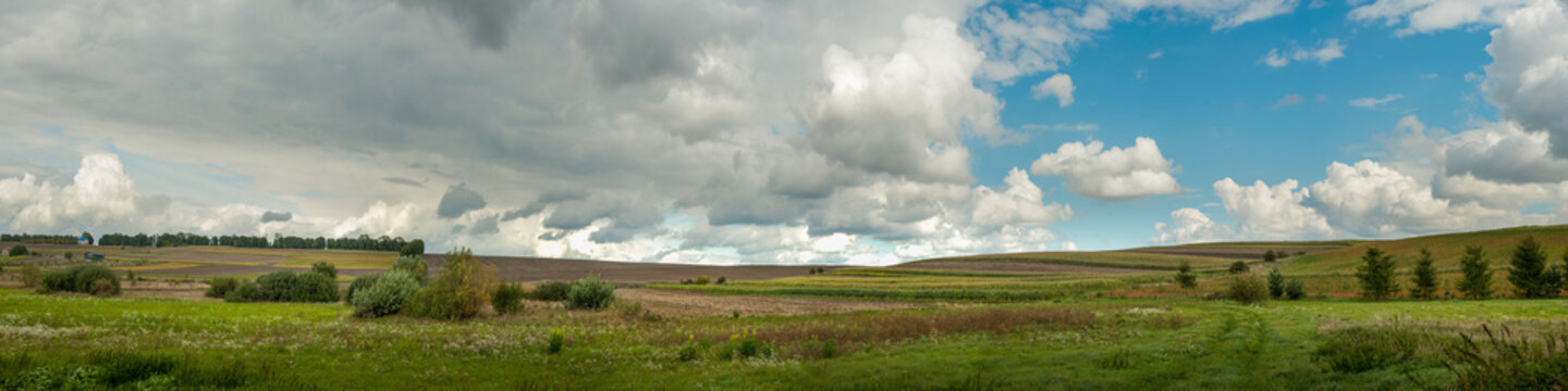 Big panoramic view of green meadows, arable fields, bushes in autumn under a cloudy sky.
