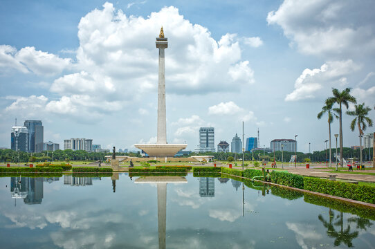 Indonesia, Jakarta &ndash; December 12, 2025: Monas tower reflected in a calm pool with green lawns and palm trees, seen from Merdeka Square under a bright cloudy sky