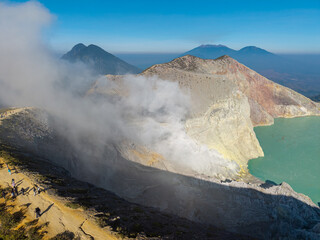 Aerial panorama of Mount Ijen turquoise crater lake, rugged volcanic rim, drifting mist, distant peaks, and visiting hikers. © Cloudyew