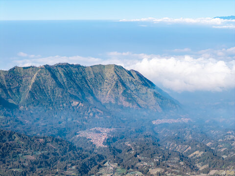 Misty peaks rise above a lush, forested valley. Clouds drift between rugged mountain ridges near Bromo