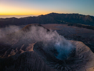 Aerial sunset over Mount Bromo, volcanic caldera, drifting mist, surrounding ridges, and distant illuminated settlements in Java. © Cloudyew