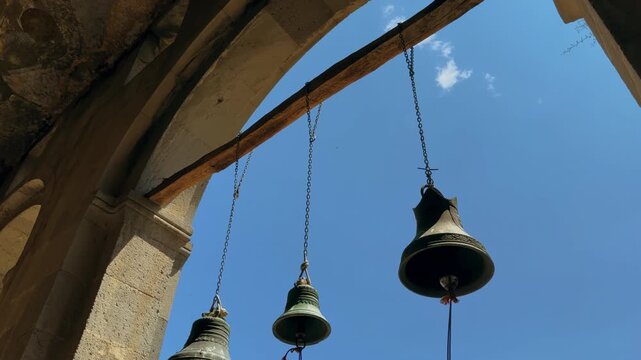 Three hanging bronze bells on metal chains beneath weathered stone arch with decorative patina against vivid blue sky in warm sunlight with dramatic shadows. Georgia