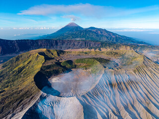Aerial daytime panorama of Mount Bromo vast crater, rugged volcanic ridges, and distant cone beneath blue skies. © Cloudyew