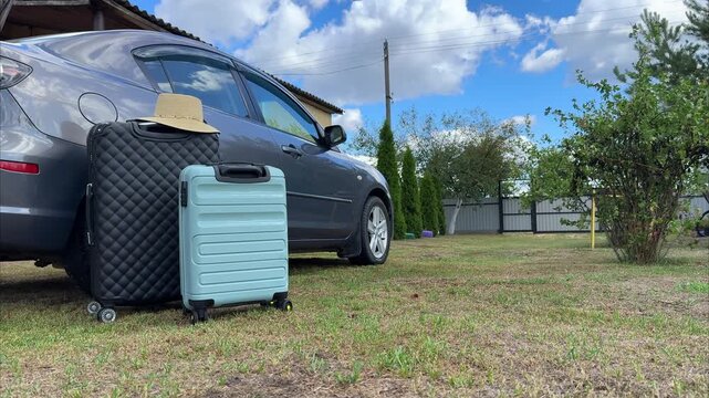 Luggage and sun hat placed beside a gray car parked on green grass under a blue sky with fluffy clouds, ready for a romantic weekend getaway