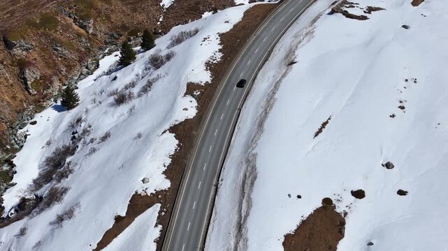 single car climbs curved mountain road cut into snowbound slope julier pass alpine crossing graubunden switzerland guardrail dry patches drone aerial view high altitude route following shot