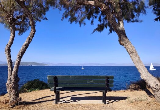 Photo of a wooden bench framed by tamarisk trees along Nikou Kazantzaki Road (O Dromos ton Logotechnon) on the island of Aegina, overlooking sailboats drifting across the the Saronic Gulf.