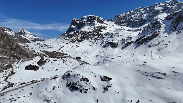 jagged snow capped peaks tower above julier pass valley floor road pylons scattered hut patchy snow graubunden switzerland drone aerial view high altitude mountain panorama clear sky