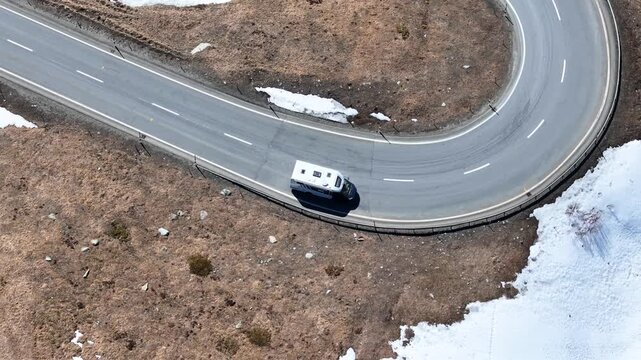 camper van completes tight hairpin loop julier pass mountain road patchy snow dry rocky terrain guardrail graubunden switzerland drone aerial view high altitude road trip alpine crossing