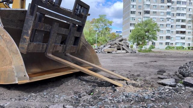 Heavy construction machinery with a bucket and wooden forks positioned on a gravel site, preparing for material handling in an urban environment