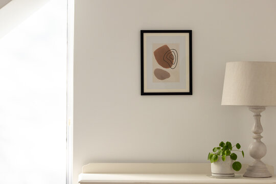 Framed abstract print is hanging on off-white wall above cream console with wooden lamp and plant