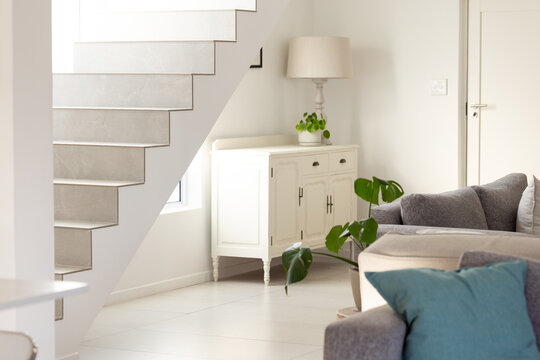 White wooden sideboard is sitting under open staircase holding lamp and trailing plant near sofa