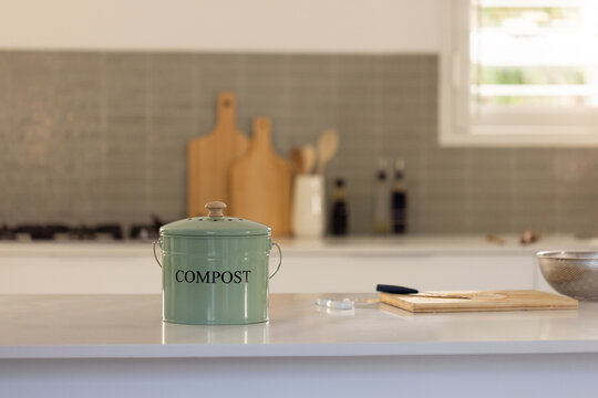 Green metal compost can is sitting on glossy white countertop island with wooden boards, colander
