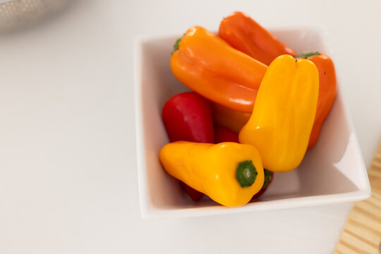 White square bowl holding glossy mini sweet peppers on smooth white table, showing shallow focus