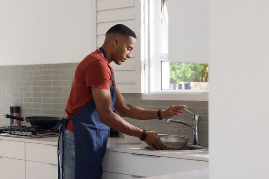 African American man washing metal colander under chrome faucet at kitchen sink wearing denim apron