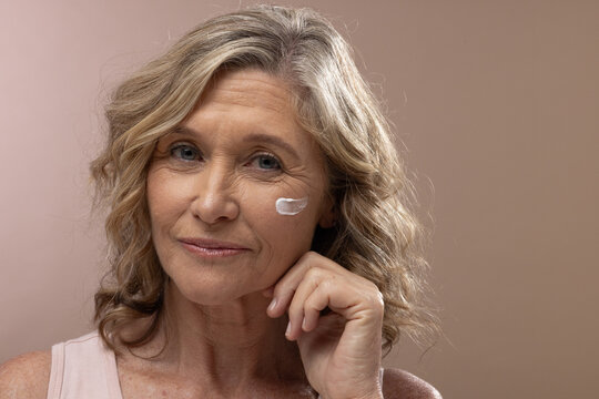 Senior woman touching face and holding chin while posing in studio with cream on right cheek