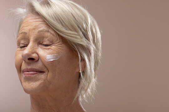 Senior woman sitting in studio, closing eyes while applying cream on right cheek with gold earring
