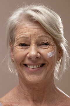 Senior woman smiling, facing camera in studio, showing cream on right cheek and wearing gold hoops