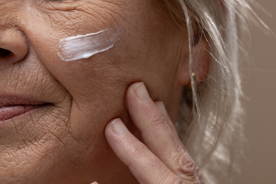 Senior woman applying white cream to cheek at sink, touching fingers and wearing gold stud earring