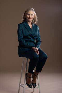 Senior female sitting on tall white stool in studio wearing teal suit and black heels