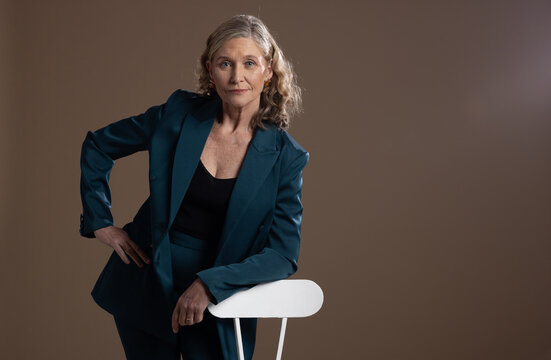 Senior woman leaning on white chair in studio wearing teal suit and black camisole, copy space