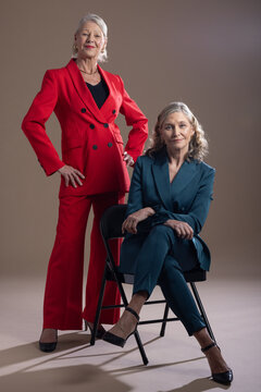 Senior female colleagues posing in studio on black folding chair, wearing double-breasted suits