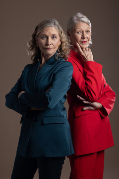 Senior women posing back-to-back in studio wearing teal and red suits with earrings