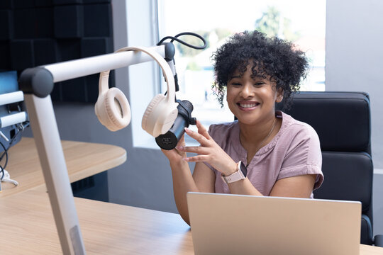 African American woman smiling and adjusting microphone while sitting at desk in studio with laptop