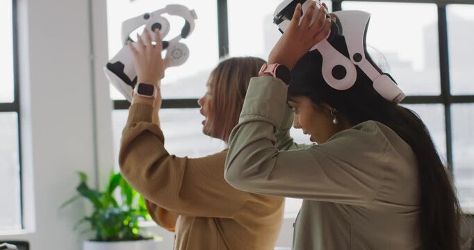 Diverse female coworkers testing white VR headsets at office by grid windows after tablet prompt