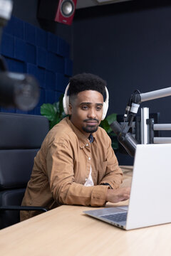 African American man recording at wood desk in studio wearing headphones, using laptop and mic
