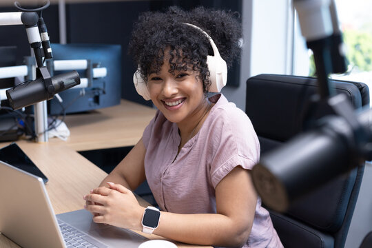 African American woman sitting at studio desk wearing white headphones, using laptop with boom mics