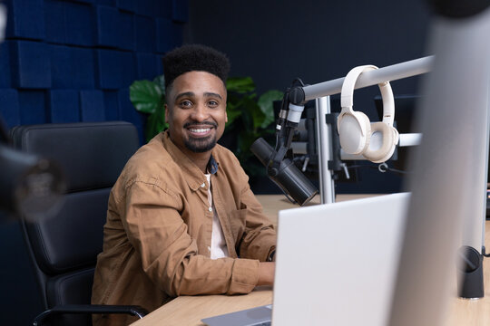 African American male smiling, sitting at studio desk wearing tan jacket, using boom mic and laptop