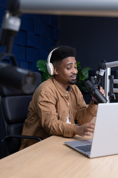 Adult African American man with white headphones talking into mic on boom using laptop in studio