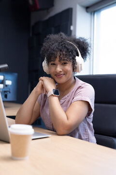 Adult African woman wearing short-sleeve blouse, headphones, podcasting in studio with laptop mic