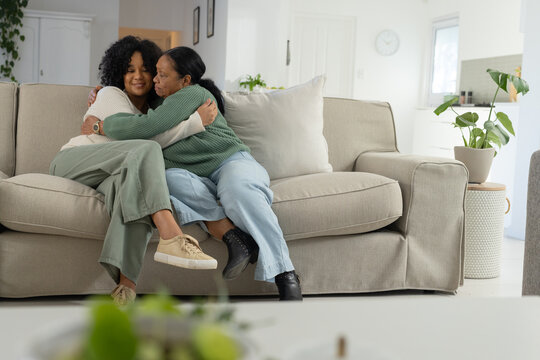 Senior African American mother and daughter sitting on beige sofa hugging with plant in living room