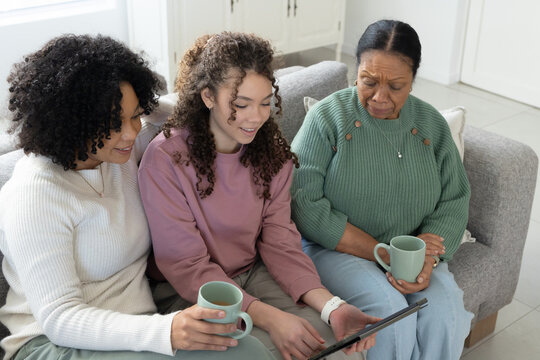 African American family sitting on sofa, in sweaters, holding light green mugs and viewing tablet