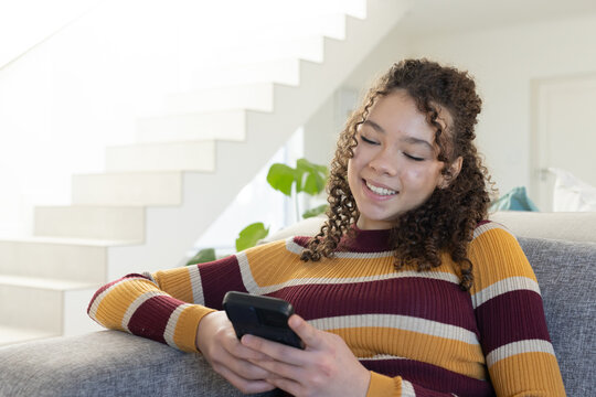 Teenage female wearing striped sweater sitting on gray sofa holding phone near stairs, copy space