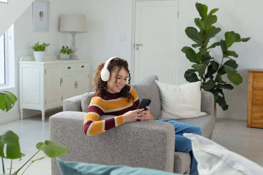 African American teen female sitting on sofa at home wearing sweater, headphones, scrolling phone