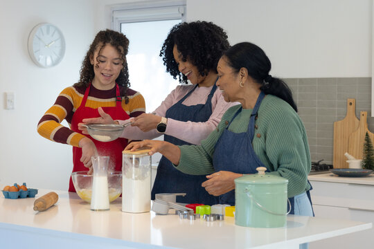 African American family gran mom teen in aprons sifting flour into mixing bowl on kitchen island