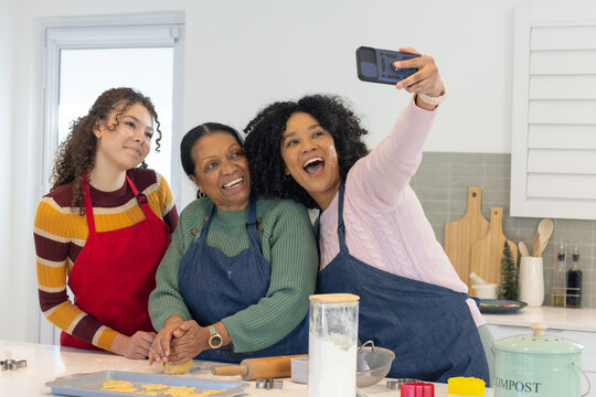 African American women baking in kitchen, senior pressing dough while teenager taking phone selfie