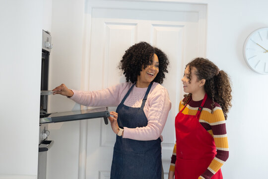 Woman and adolescent female standing in home kitchen holding oven door pulling tray wearing aprons