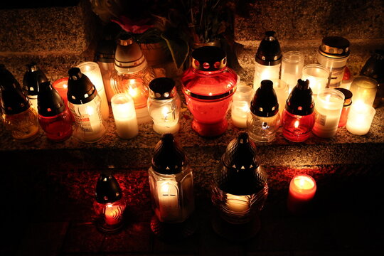 Candles lit in memory of deceased relatives during All Souls' Day. Many lit candles rest in glass and metal holders. Red and white flames illuminate dark setting. Candles create soft, reverent atmosph