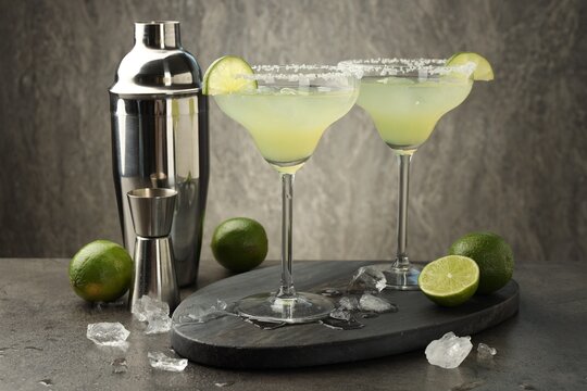 Refreshing margarita cocktail in glasses, limes, shaker and jigger on grey table, closeup