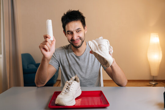 Vertical portrait of cheerful young man holding white sneaker and shoe cleaner bottle, preparing to clean footwear at home, demonstrating process of refreshing shoes and maintaining appearance.