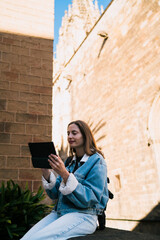 Caucasian female student holding digital tablet computer sitting near stone architecture, listening to audiobooks and managing mobile banking apps for flexible lifestyle © BullRun