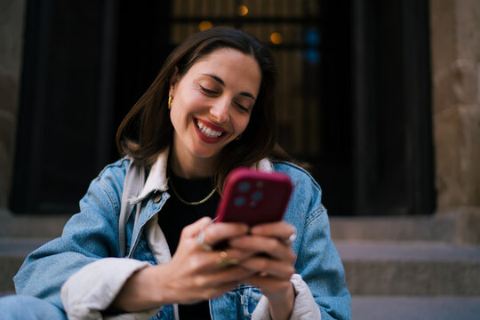 Radiant Caucasian female student laughing while looking down at smart phone sitting on steps, enjoying cybersecurity safe social messaging and dynamic modern digital connection