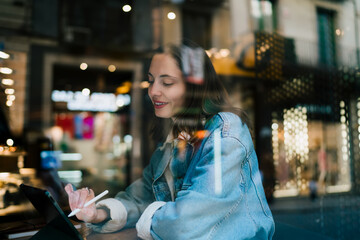 Through window shot of focused Caucasian female student using touch stylus pen on tablet computer in cafe, managing e-commerce retail store products and online shopping orders in city © BullRun