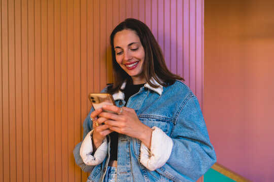 Smiling Caucasian female student holding smartphone leaning against pink wall, exploring e-commerce mobile retail store deals processing instant checkout and modern shopping apps in city